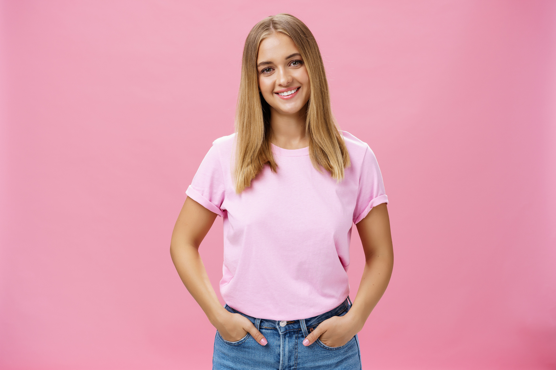 Charming Pleasant Woman with Chubby Face and Fair Hair in Casual T-Shirt Smiling Broadly Holding Hands in Pockets Being Nice and Friendly Gazing Carefree and Calm at Camera over Pink Background