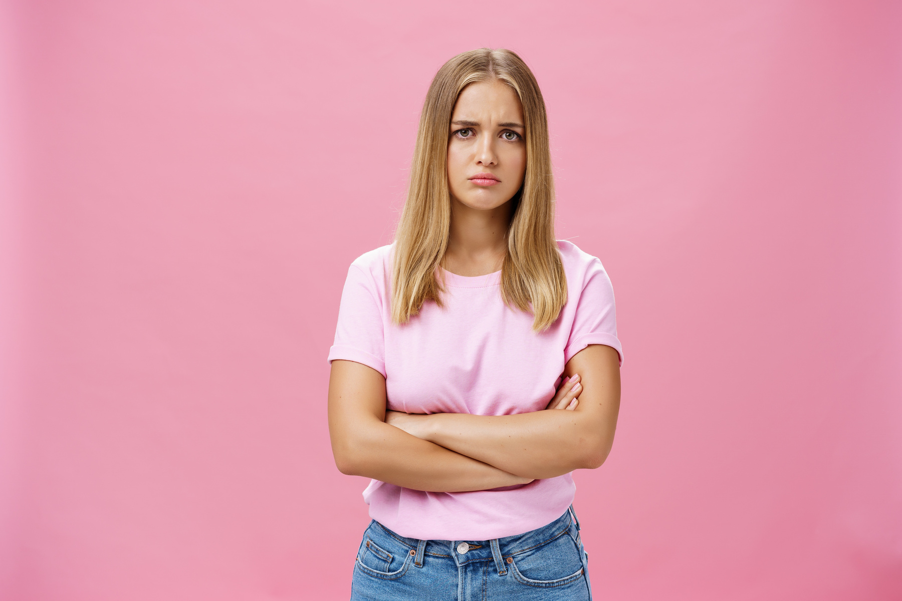 Sad Envy and Disappointed Gloomy Cute Young Woman with Fair Hair in Casual T-Shirt Crossing Arms against Chest in Upset Gesture Pursing Lips and Frowning Looking Concerned and Sad over Pink Wall